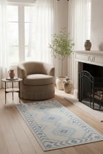 A rectangular area rug with a light blue and off-white geometric pattern is styled on a light wood floor in a bright, modern living room next to a beige armchair, a small side table, and a fireplace.