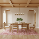 A dining room scene with a light wood oval table and six woven cane chairs with off-white seat cushions, arranged on a pink patterned area rug. A brass chandelier hangs above, and the room has warm wood ceiling beams, cream walls, and arched openings in the background.