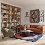 A styled living room interior featuring a rust-orange sofa, a green armchair, a round glass-top coffee table, a blue rug with orange and cream circular motifs, a large light wood bookcase filled with books and decor, and framed wall art above the sofa.