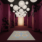 A long hallway with dark walls, black splatter-like patterns, and many white hanging paper lantern lights. A light blue rectangular rug with floral medallions lies on the floor in the foreground.