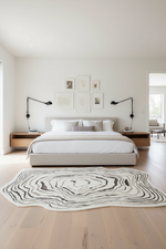 A modern bedroom featuring a low-profile platform bed with white bedding, a large organic-shaped black and white abstract patterned rug on light wood flooring, and black adjustable wall sconces on wooden nightstands.