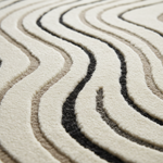 Close-up of a plush textured rug with wavy, concentric wavy patterns in cream, beige, taupe, and black.