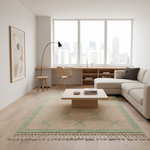 A modern living room with a light beige sectional sofa, a low wooden coffee table, and a large beige area rug with green fringe and geometric patterns. The room features light wood floors, a minimalist desk by a large window, and a framed abstract art print on the wall.