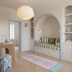 A beautifully styled interior room featuring a built-in daybed with green and white striped cushions under a large arched niche. The room has light wood floors, a pink and blue patterned runner rug, a large white pleated pendant light, and built-in shelving with decorative items.