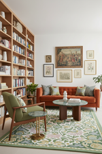 A styled living room featuring a rust-orange tufted sofa, a green armchair, a large floral green and cream area rug, and a tall wooden bookshelf filled with books against a white wall with framed art.
