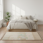 A styled bedroom scene with a bed dressed in cream bedding and a textured gray throw, set in a bright room with a light beige woven area rug in the foreground, plants, books, and a side table with a lamp.