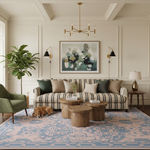 A beautifully styled living room featuring a striped sofa with green and beige pillows, a blue and pink patterned area rug, rustic wooden coffee tables, a green armchair, and a sleeping dog on the floor. A large framed abstract painting hangs on the cream-colored paneled wall.