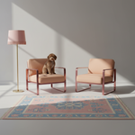Two blush pink armchairs with rose gold metal frames are arranged in a minimalist room with a small brown dog sitting on one chair. A matching pink floor lamp stands on the left, and a blue and pink patterned rug covers the floor.