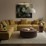 A modern living room featuring a curved mustard yellow sectional sofa, a round dark brown marble coffee table, a patterned area rug, and a large abstract art print on the wall.