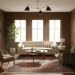 A beautifully styled living room featuring a cream-colored sofa as the central focus, set on a traditional patterned area rug. The room has warm wood-paneled walls, three tall windows, a brass three-arm chandelier, a green plaid armchair, and a potted plant.