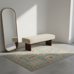 A styled interior scene with a cream boucle bench on a patterned light blue and beige rug, beside a tall floor mirror with a gold frame against a white wall.