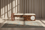 A modern wooden bench with a white cushion sits on a patterned area rug in a sunlit room with beige curtains. The bench features rich wood grain panels and a round white marble inlay, with a matching marble side table beside it.