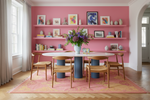 Dining room with pink rug, pink walls, shelves, and table, featuring a vase of flowers and books.