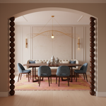 A styled dining room interior viewed through a rounded archway with dark wooden posts. The room features a round dark wood dining table, four blue upholstered chairs, a cream sofa, a pink patterned rug, and a gold wire pendant light.
