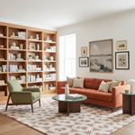 A styled living room interior featuring a terracotta orange fabric sofa with striped pillows, a green armchair, a round glass-top coffee table, a patterned rug, and a large built-in wooden bookshelf against the wall.