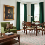 A styled interior room with a large framed portrait on the wall, deep green curtains, a green armchair with a leopard-print pillow, a wooden dining table and chairs, a patterned rug, and a coffee table in the foreground.