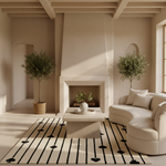 A modern, minimalist living room featuring a curved cream sofa, a low concrete coffee table, a cream rug with black geometric patterns, and a central fireplace. Two potted olive trees stand on either side of the fireplace.