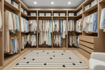 A spacious walk-in closet with light wood cabinetry, shelves, and hanging garments in soft neutral and pastel tones. A striped rug with black heart motifs lies on the floor, and part of a white chair is visible in the lower right corner.