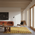 A yellow area rug with black polka dots and pink circles is styled in a modern, sunlit living room with a brown leather sofa, wooden ceiling beams, and a black accent chair.
