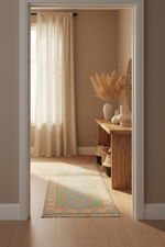 A long runner rug with a light green, beige, and orange floral pattern is styled on a light wood floor in a doorway, leading to a sunlit room with cream curtains and a wooden console table.