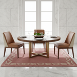 A round wooden dining table set with four brown leather chairs, place settings, wine glasses, and a bowl of fruit, arranged on a pink patterned area rug in front of a white marble-tiled wall and window.