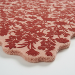 A close-up of a plush, textured area rug with a scalloped edge, featuring a dusty rose pink background with a raised rust-red floral pattern.