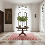 A round wooden pedestal table centered in a bright room with white curtains, a large arched window, a vase of greenery, and a pink-and-white checkered rug beneath it.
