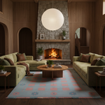 A cozy retro-style living room with two olive green sectional sofas facing a stone fireplace, a round wooden coffee table, a light blue and coral checkered rug, and a large white paper lantern hanging above the fireplace.