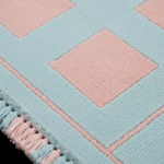 Close-up of a textured rug with a geometric diamond pattern in pastel pink and light blue, featuring a tassel fringe along one edge.