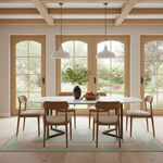 A modern dining room with a round white table, six wooden chairs with cream upholstery, a green patterned rug, and large arched windows looking out to a garden. A black vase with a leafy plant and a bowl of fruit sit on the table, with two white pendant lights hanging above.