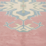 A close-up of a pink rug with a traditional geometric pattern in cream, light blue, and pale green, shown in a shallow depth of field.