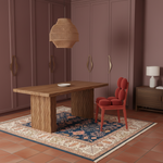 A styled interior scene showing a wooden dining table with textured block legs, a coral-red upholstered chair, a woven pendant light, a patterned rug, and mauve-toned wardrobes in the background.