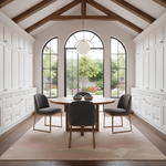 A modern dining room with a round wooden table, three gray upholstered chairs with gold legs, a pink patterned area rug, and tall white cabinets on both sides under a high vaulted ceiling with exposed wood beams.