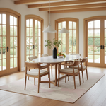 A dining room scene with a round white table and six light wood chairs with cream cushions, arranged on a pale pink rug in a bright room with large arched glass doors and hanging pendant lights.