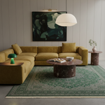 A modern living room setup featuring a mustard yellow sectional sofa, a green patterned area rug, a round marble-top coffee table, and a large abstract art print on the wall.