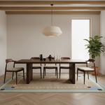 A modern dining room setup featuring a dark wood rectangular table and four matching chairs with light beige upholstered seats, arranged on a patterned area rug. A large potted fiddle-leaf fig plant stands in the corner next to a tall window, and a ribbed white pendant light hangs above the table.