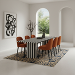 A modern dining room scene featuring a rectangular table with bold black and white vertical stripes, surrounded by six orange upholstered chairs on a patterned beige and navy rug. The room has white walls, arched windows looking out to green trees, and a large abstract black-and-white framed print on the left wall.