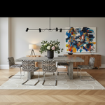A modern dining room scene with a rectangular gray stone table, six zebra-print upholstered chairs, a black pendant light, a vase of flowers, a sideboard, and a large abstract painting on the wall.