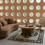 A styled living room scene featuring a curved tan sofa, a matching accent chair, a round wooden coffee table, and a patterned rug in front of a terracotta wall with circular cutouts.