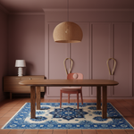 A styled dining room scene featuring a rectangular wooden dining table on a blue and cream patterned area rug, with a pink chair tucked underneath. A large woven pendant light hangs from the ceiling, and the background consists of dusty rose paneled walls and built-in cabinetry.