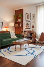 A styled living room featuring a green tufted sofa, a wooden coffee table, a cream rug with blue abstract lines, a brown leather chair, and a slatted wood accent wall with framed art.