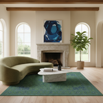 A styled living room interior featuring a curved olive green sofa, a white marble coffee table, a green patterned area rug, a stone fireplace, and a large blue abstract canvas painting on the wall.