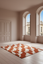 A rectangular area rug with an abstract organic pattern in terracotta orange and cream, displayed on a light wood plank floor in a modern, minimalist room with beige walls and arched windows.