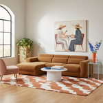 A styled living room featuring a large rust-brown sectional sofa as the main product, placed on a patterned rug with a marble coffee table, a pink armchair, plants, and wall art in a bright neutral interior.