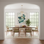 A styled dining room interior with a wooden dining table, six woven and upholstered chairs, a hanging brass chandelier, large black-framed windows, a colorful abstract painting, and a potted plant, viewed through a white arched doorway.