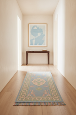 A long runner rug with a blue and beige medallion pattern and fringe, styled on a light wood floor in a minimalist hallway with a console table and abstract art in the background.