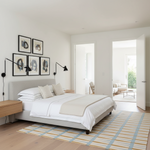 A modern bedroom featuring a light gray upholstered bed with white bedding, a beige throw blanket, and a striped blue, yellow, and white rug on a light wood floor. The wall above the bed has four black-framed abstract prints.