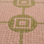 Close-up of a textured woven rug with a geometric pattern in sage green on a dusty pink background.