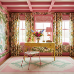 A vibrant interior room featuring a yellow patterned desk, a pink metal chair, and matching pink and multicolor floral wallpaper and curtains. The room is decorated with a pink lamp, a floral vase, and a geometric pink and green rug.