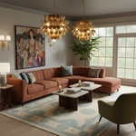 A styled living room featuring a rust-orange sectional sofa, two ornate gold leaf chandeliers, a blue and beige patterned area rug, and a large framed painting on the wall.